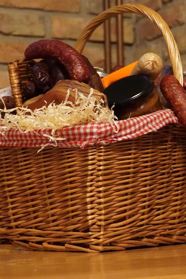 Proutěný uzenářský koš s klobásami, uzeninami a sklenicemi na kostkovaném ubrusu